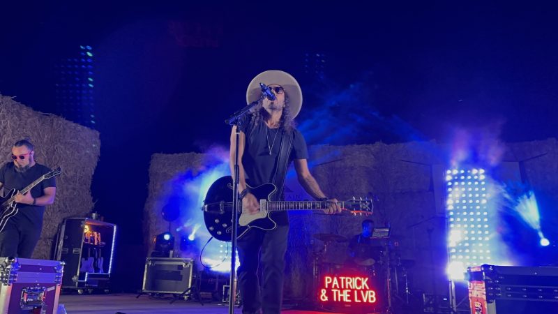 Patrick in a cream cowboy hat laughs alongside a bandmate in red sunglasses as they share a guitar in front of stacked hay bales.