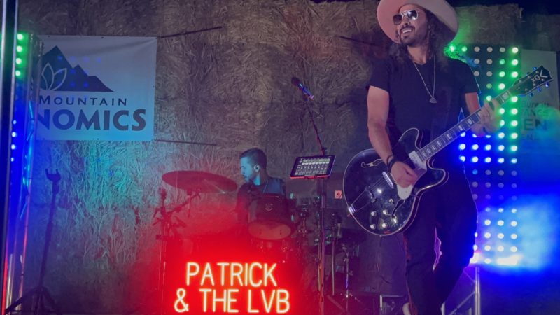 Patrick in a cream cowboy hat and sunglasses, black guitar slung across his chest, the red neon Patrick & The LVB sign glowing on the hay-bale stage behind him.