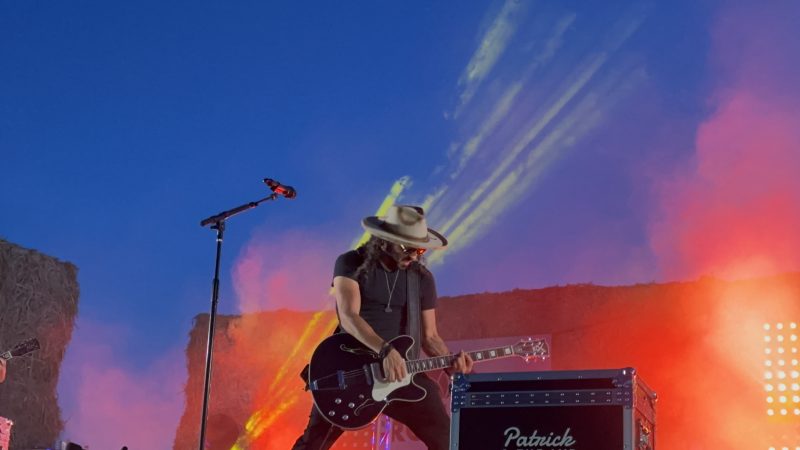 Patrick crouches over his black guitar at blue hour, the sky above streaked with sunset clouds and vapor trails, red and orange light spilling from the hay-bale stage.