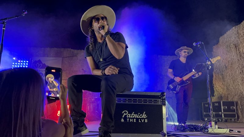 Patrick sings while seated on a Patrick & The LVB road case as a fan in the front row holds up her phone to film him, her screen showing his image.