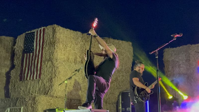 Patrick raises his guitar overhead facing away from the camera, a full American flag hanging on a wall of golden hay bales to his left and a bandmate playing guitar to his right.
