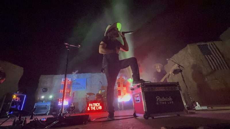 Low-angle silhouette of Patrick with one foot up on a road case, microphone to his mouth, a red-lit American flag and hay bales filling the right side of the frame.