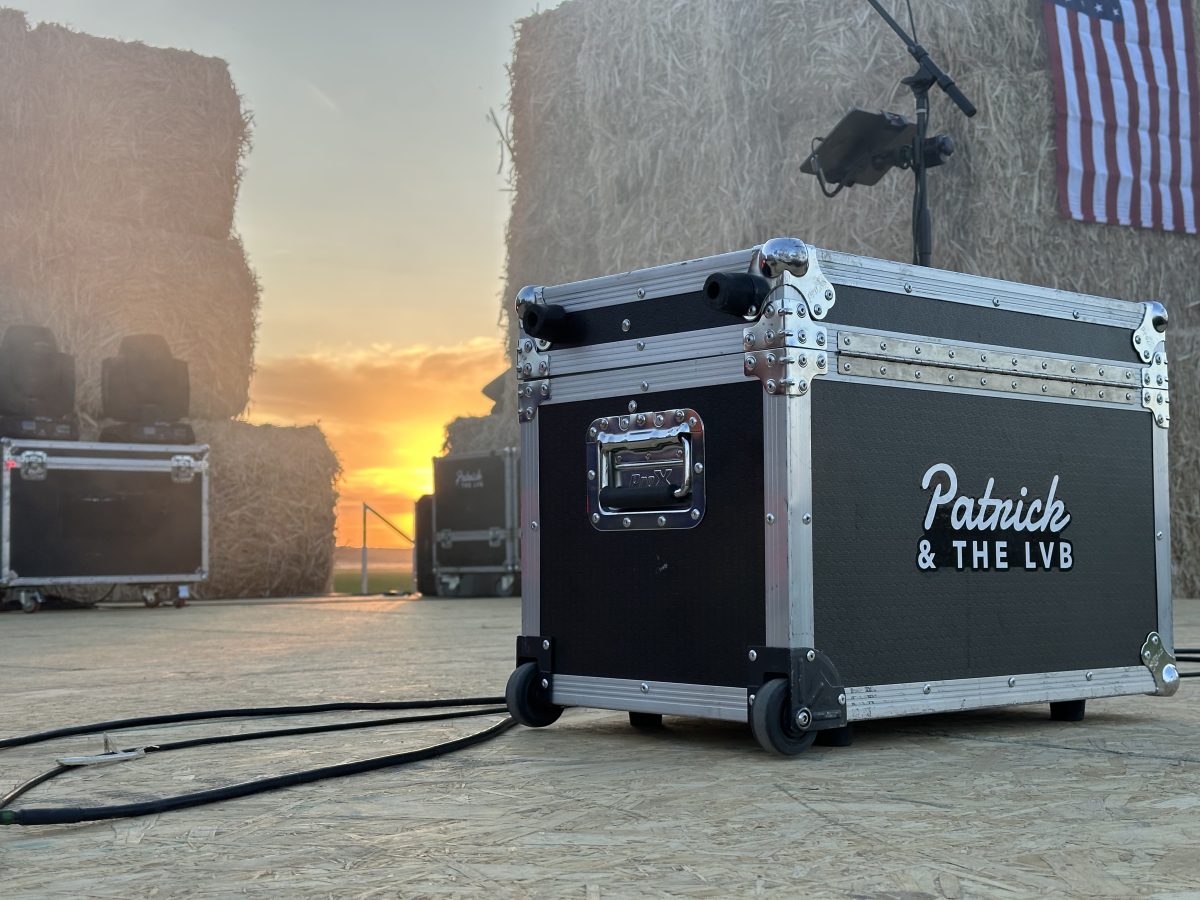 A Patrick & The LVB road case on an empty plywood stage at sunset, framed by hay-bale walls, a mic stand, and an American flag, with the sun setting over the Idaho horizon.