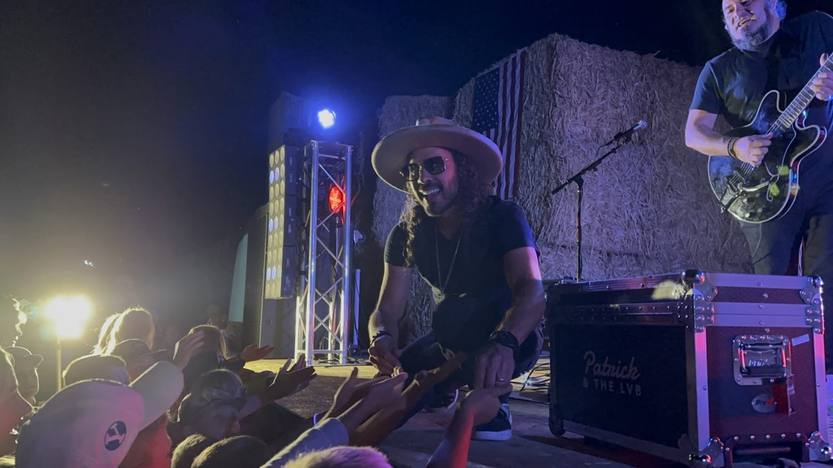 Patrick crouches at the edge of the stage smiling down as small hands reach up from the front row, the American flag on the hay bales lit blue behind him.